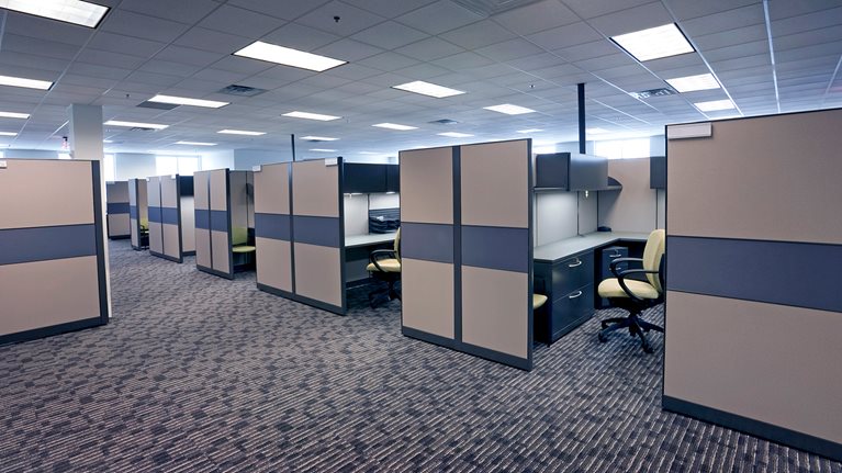 An office floor filled with beige-colored, carpeted flooring and rows of cubicles that all look the same. Each cubicle contains a desk, chair, and is lit by overhead fluorescent lighting.