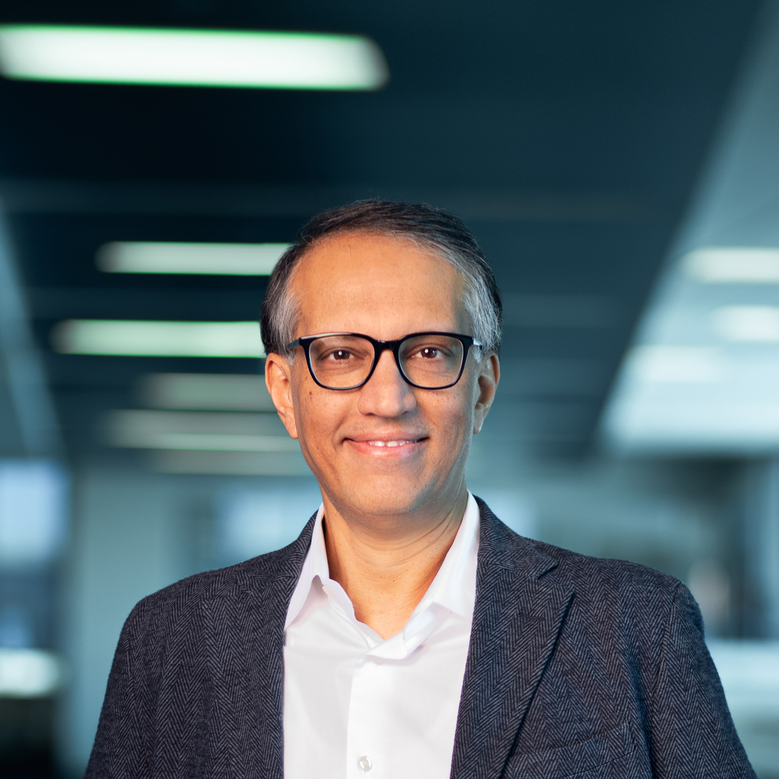Portrait of a smiling Vipul Chawla shot against a blurred out office setting. Chawla is wearing glasses and a dark gray sport coat and button down shirt.