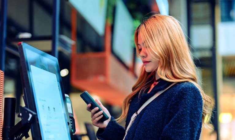 Women in casual clothing holding up her cell phone as she uses a smart checkout screen in a department store.