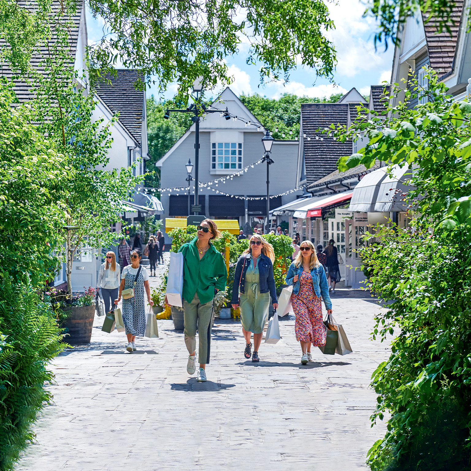Shoppers carrying shopping bags walk outdoors through Bicester Village. 