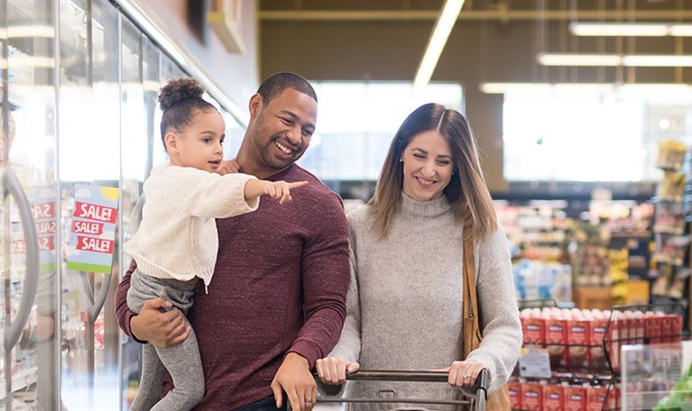 Dad holds his pre-school age daughter at the grocery. Mom is next to him pushing the cart. They are both chuckling as they stroll through the refrigerated dairy section.