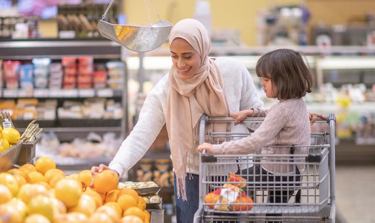 A Muslim mother goes grocery shopping with her daughter. Her daughter is sitting on the grocery cart while she picks through some fresh produce.