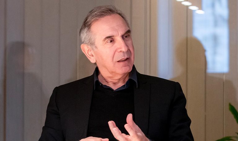 Carlo Capasa at a table, wearing a dark blazer and engaging in animated conversation, his hands gesturing expressively.