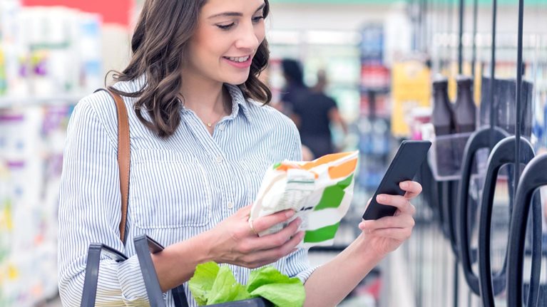 Confident young Hispanic woman holds smart phone as she reads a nutrition label on a bag of frozen vegetables. She is holding a shopping basket filled with healthy foods.