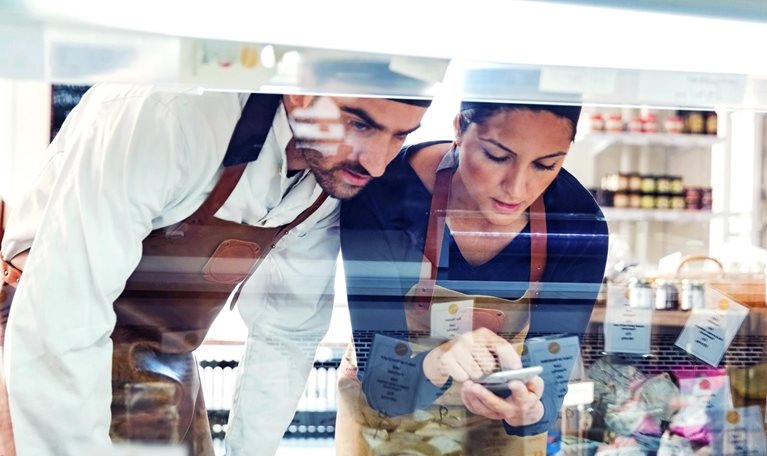Male and female business owners wearing smocks using smart phone while examining display cabinet in a grocery store