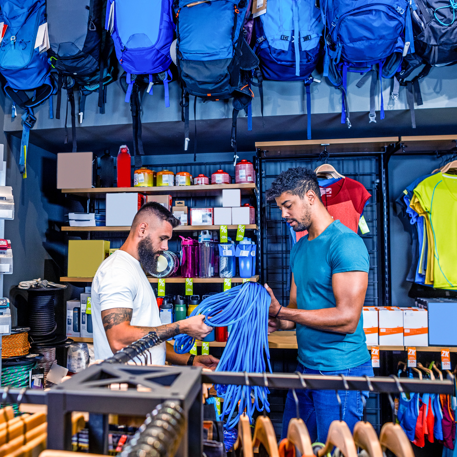 Two men examining a coil of blue climbing rope in an outdoor sports store, surrounded by shelves of gear and clothing. The shelves are stocked with various items, including water bottles, camping supplies, and colorful shirts.