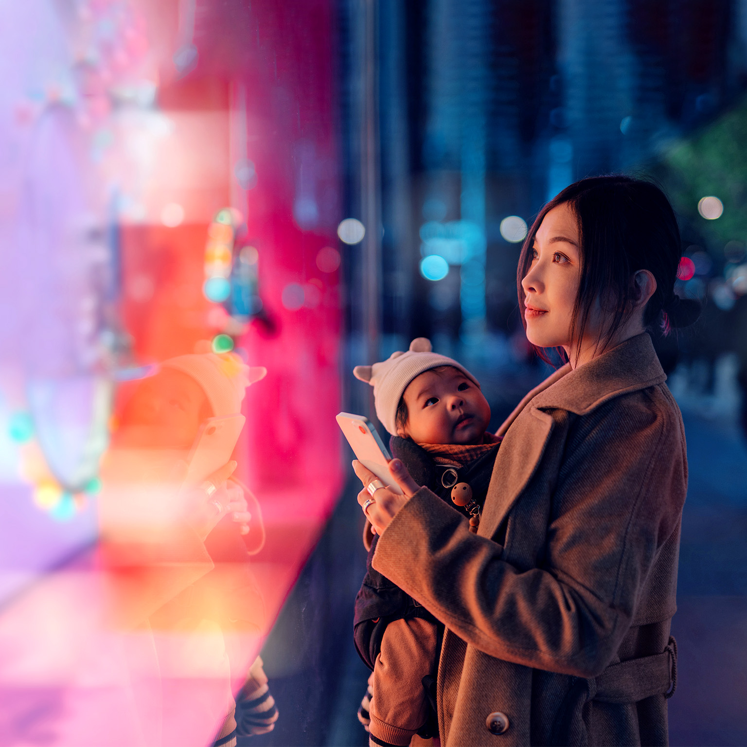 Mother carrying her baby and using a smartphone while looking at a brightly lit shop window display.