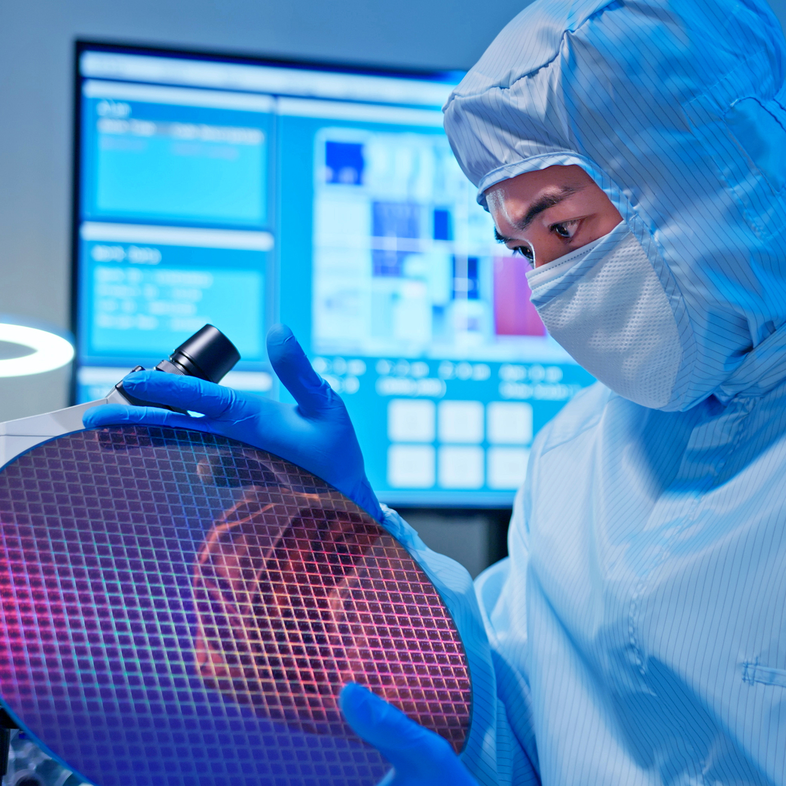 Asian male technician in sterile coverall holds wafer that reflects many different colors with gloves and check it at semiconductor manufacturing plant.