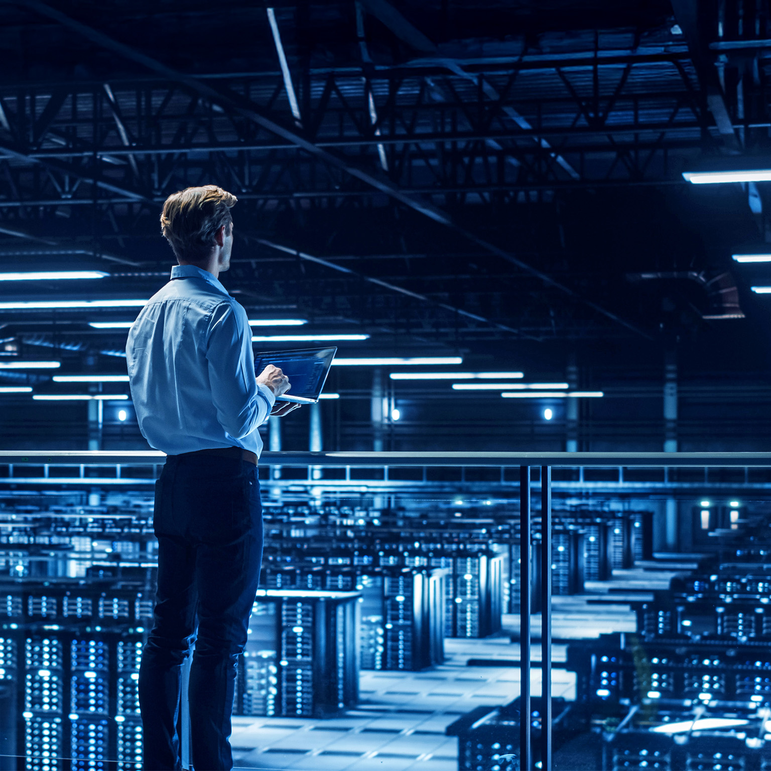 A person standing on an elevated walkway in a large, blue-lit data center, holding a laptop and overlooking rows of server racks.