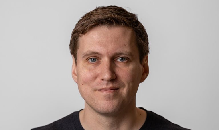 Philip Johnston with short brown hair and blue eyes looks directly at the camera with a slight, relaxed smile. He wears a dark shirt and is set against a plain, light-colored background.