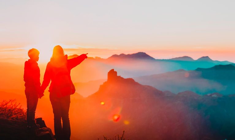 mother holding hand of son while pointing over canyon rim, sun setting in background - photo