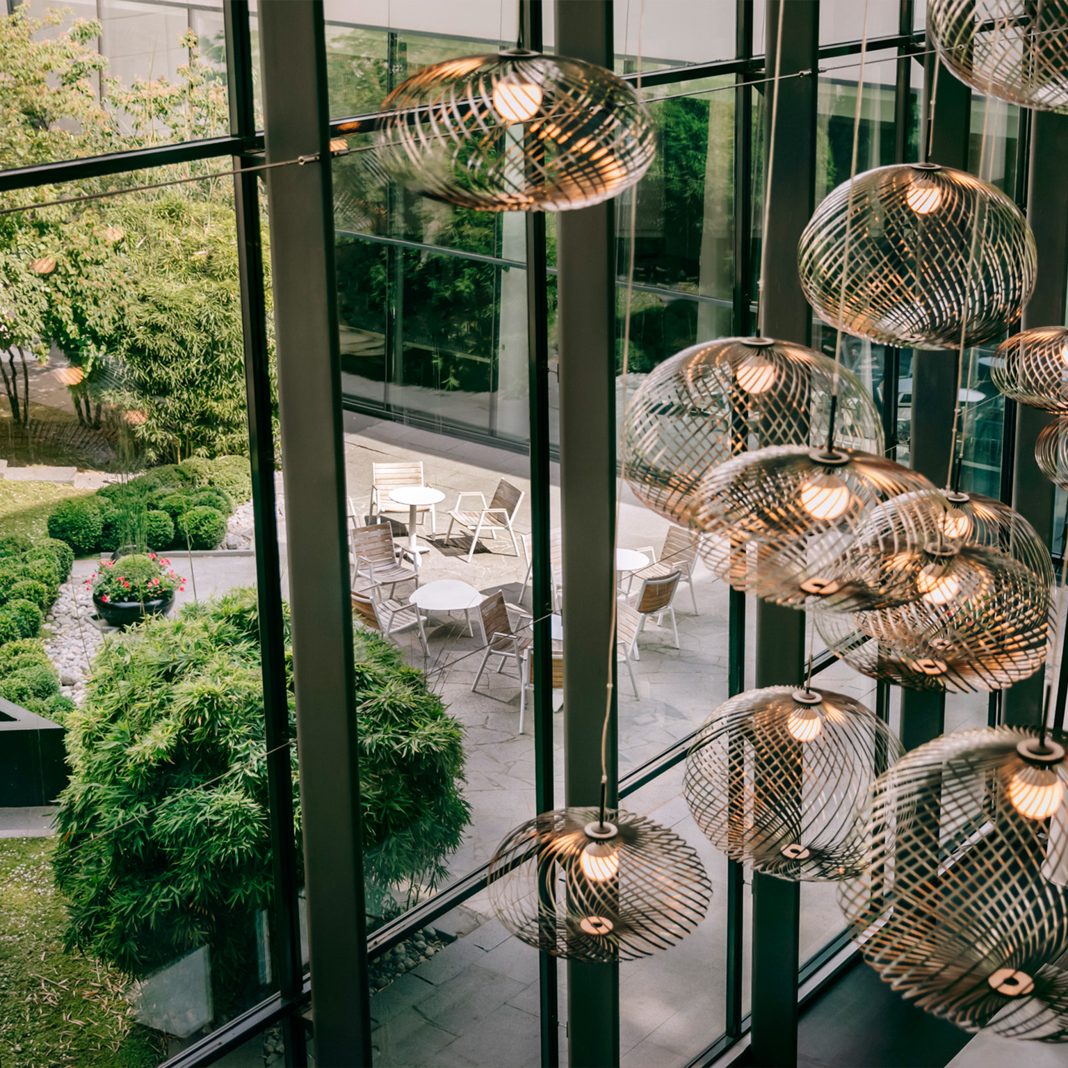 View from inside an Accor hotel through a modern glass wall and golden light fixtures out onto patio seating surrounded by greenery.
