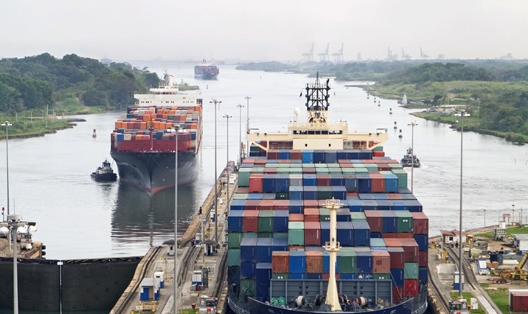 Cargo ships assisted by tugboats, entering the Panama Canal at Gatun Locks on the Atlantic side. The ships are fully loaded with cargo heading west towards the Pacific.