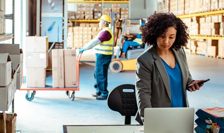 photo warehouse with woman looking at laptop while men move boxes in background
