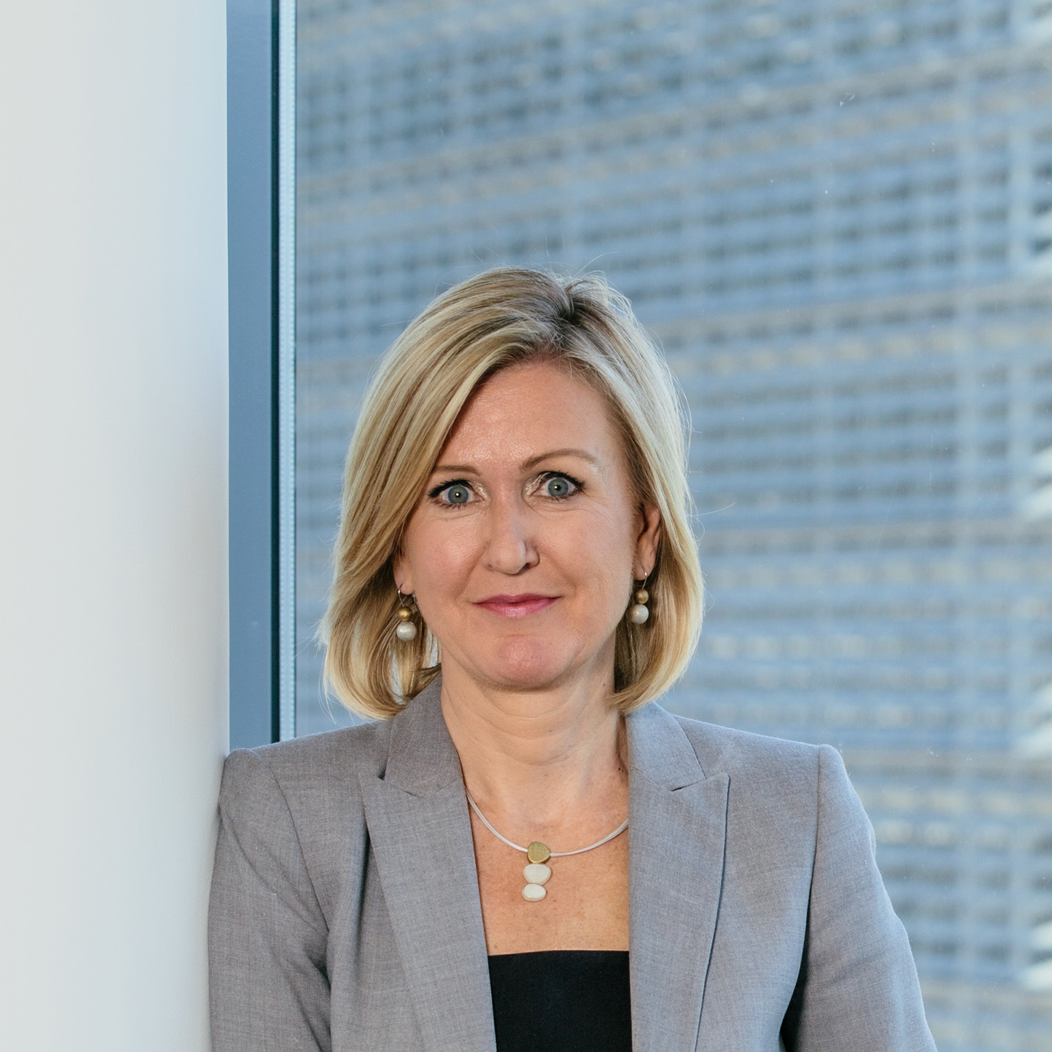 Portrait of Ann Mettler leaning against a pillar with large windows behind her in a modern office building.
