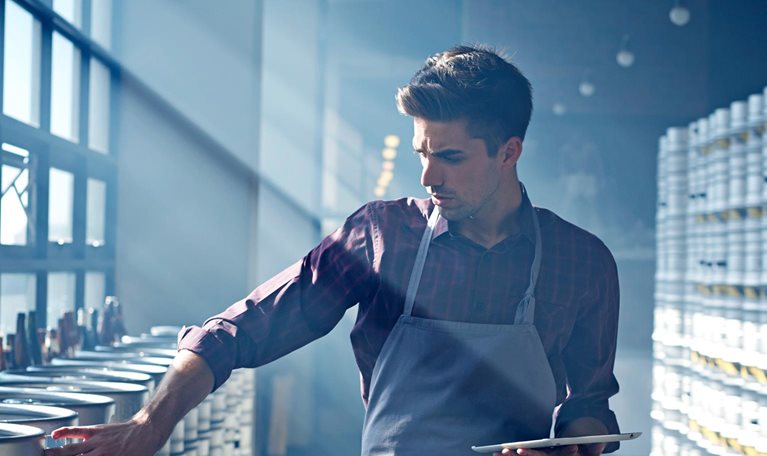 Quality control specialist in apron inspecting beer barrels in brewery, holding digital tablet in hands