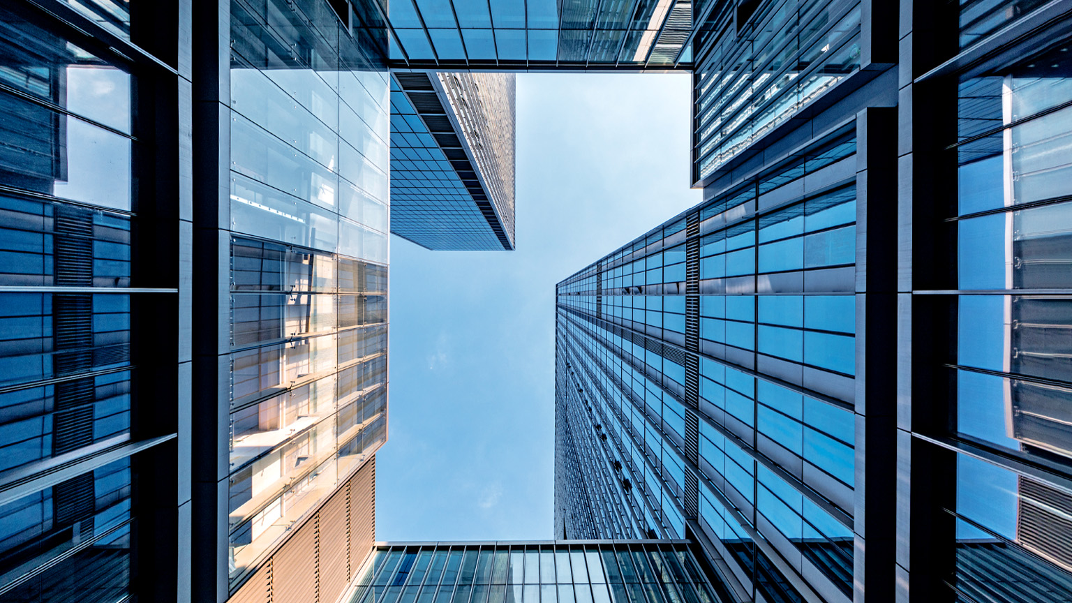 A dramatic upward view between modern glass skyscrapers, their reflective blue surfaces forming a geometric frame around the sky. 