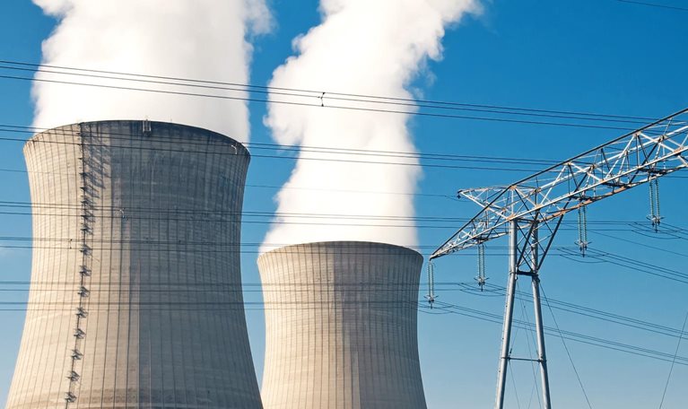 Two large cooling towers at a power plant releasing thick white steam into a clear blue sky, with high-voltage transmission lines and electrical towers in the foreground.
