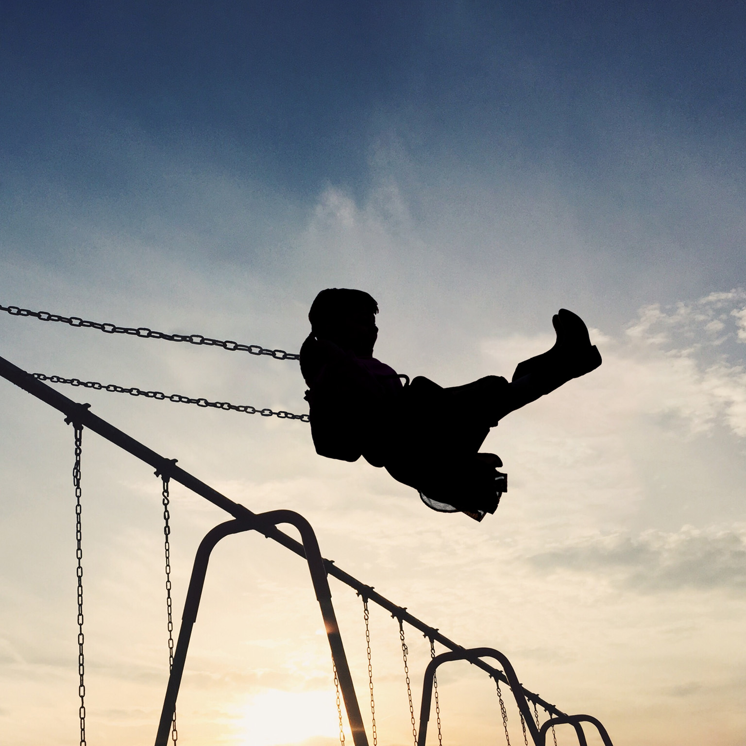 A silhouetted child soars through the air on a swing set. The setting sun casts a warm glow on the scene, creating a quiet and nostalgic atmosphere.