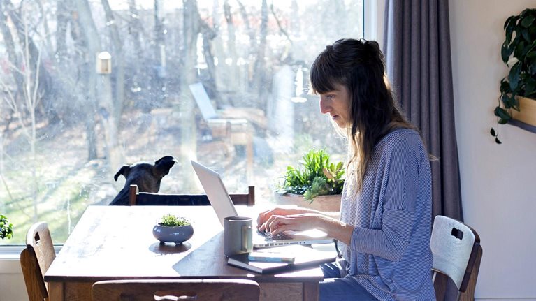 A woman working on a laptop at her dining room table in a suburban home. A dog is seen peering out the adjacent window onto a wooded yard.