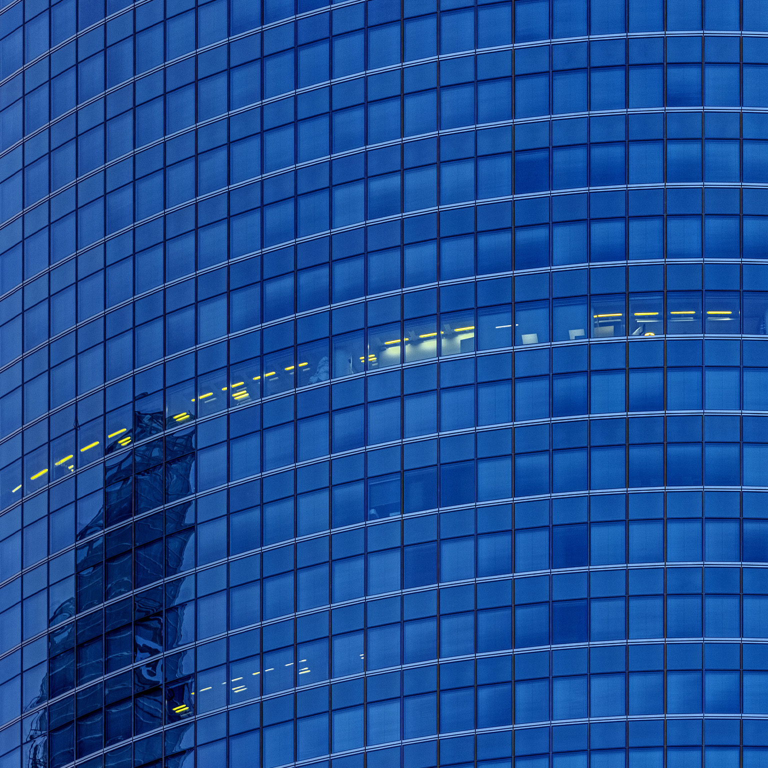 A detail shot of a glass façade skyscraper. All the floors appear dark except for a smattering of offices which have their lights on.