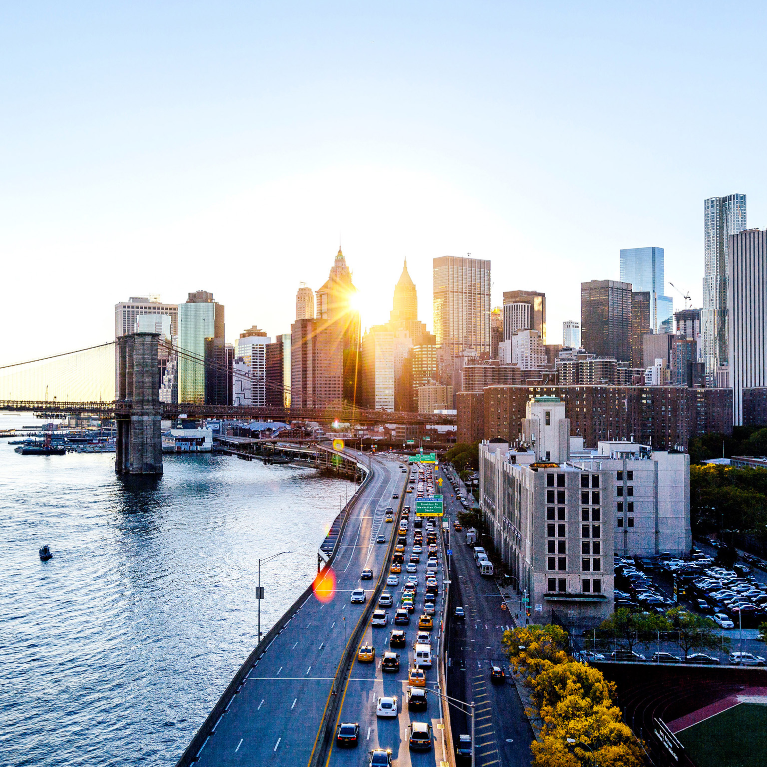 Sunset over lower Manhattan with the Lower East Side in the foreground and the Financial District in the background. The Brooklyn Bridge intersects the scene with a busy FDR Drive framing the cityscape.