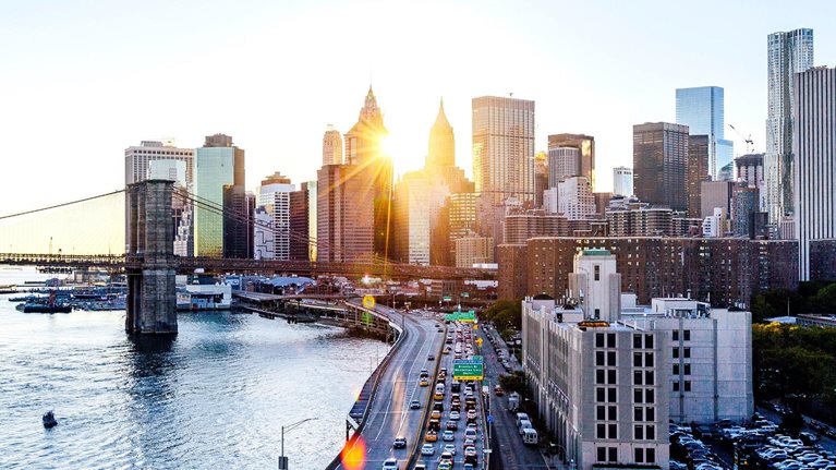 Sunset over lower Manhattan with the Lower East Side in the foreground and the Financial District in the background. The Brooklyn Bridge intersects the scene with a busy FDR Drive framing the cityscape.