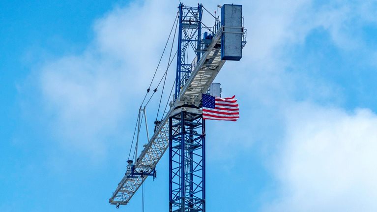 An American flag attached near the top of a tall construction crane waves in the wind. The crane rises against a cloudy blue sky, with its long arm extending outward.