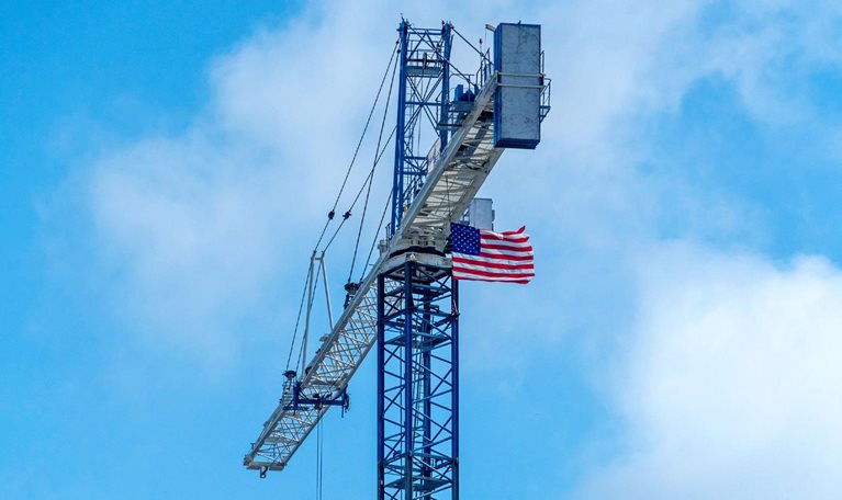 An American flag attached near the top of a tall construction crane waves in the wind. The crane rises against a cloudy blue sky, with its long arm extending outward.