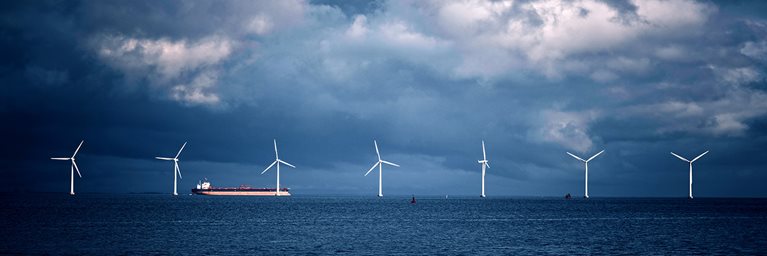 Wide view image of wind turbines in the ocean with turbulent cloudy skies and an oil tanker going by in the distance.