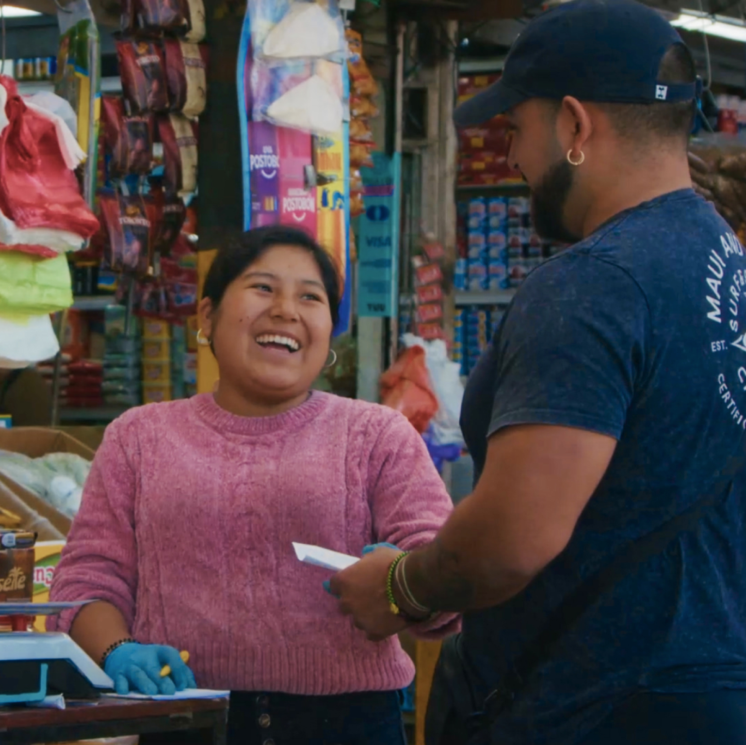 Photograph of a small grocery store checkout area showing a cashier wearing a pink sweater and gloves interacting with a customer in a blue shirt and cap. Shelves behind the cashier are stocked with various packaged food items, including biscuits, cereals, and snacks, highlighting a typical local market setting.