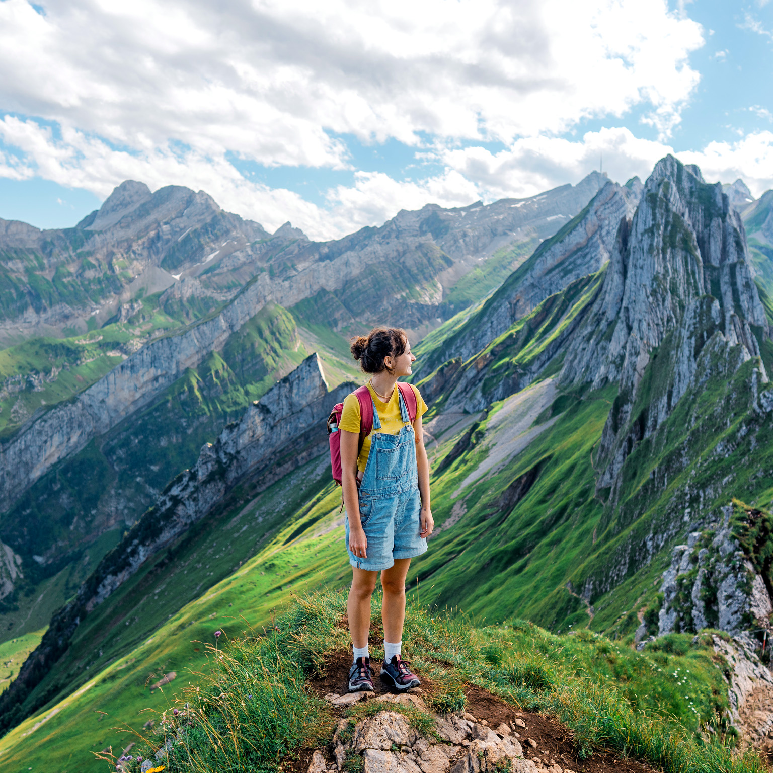 A woman wearing hiking clothes and a red backpack stands on a grassy mountain ridge surrounded by the steep rocky peaks and green valleys of the Swiss Alps.