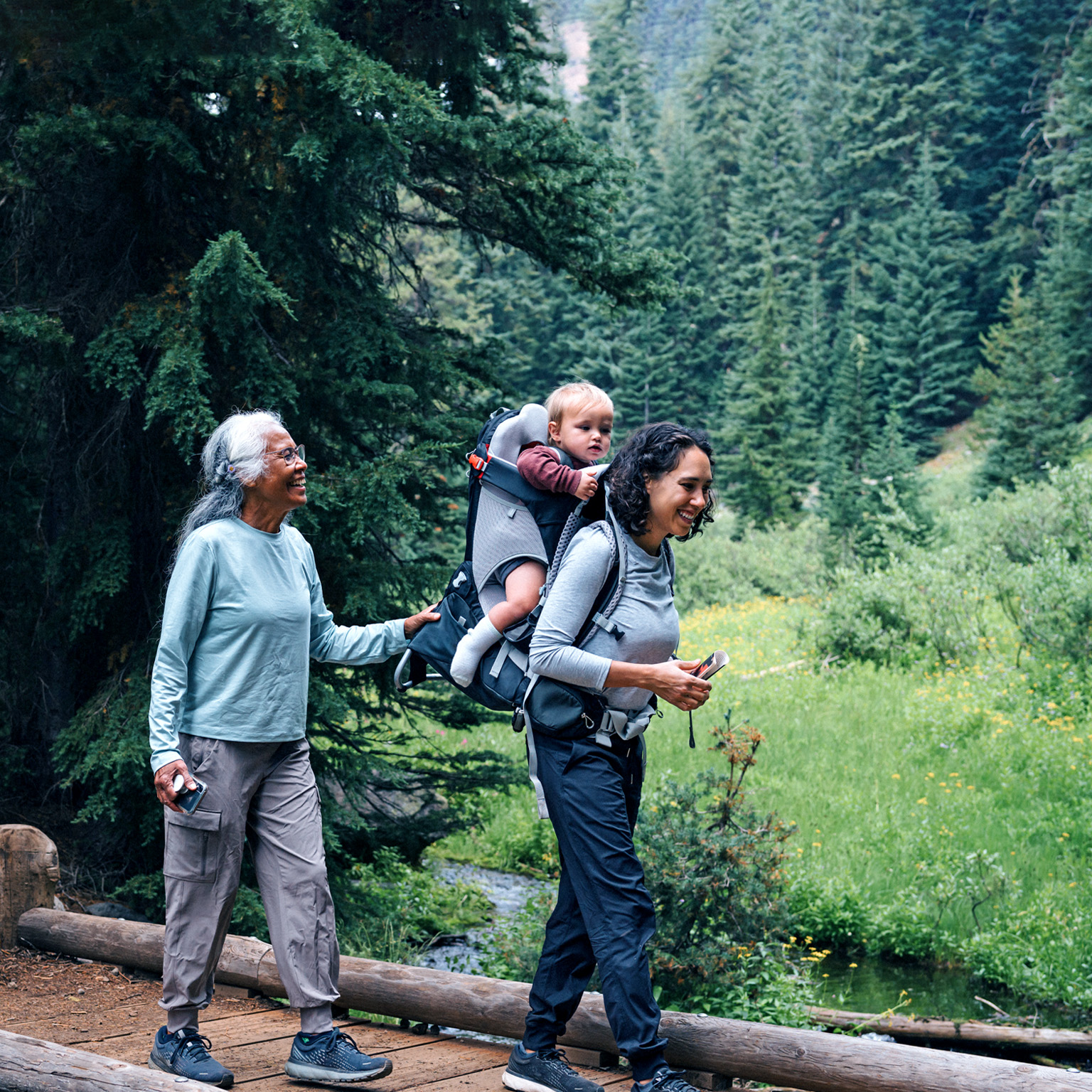 A smiling family, composed of a grandmother, grandfather, mother with a baby in a carrier on her back, and a young girl, treks through a forest, crossing a wooden bridge over a small stream. The grandfather assists the girl as they walk.