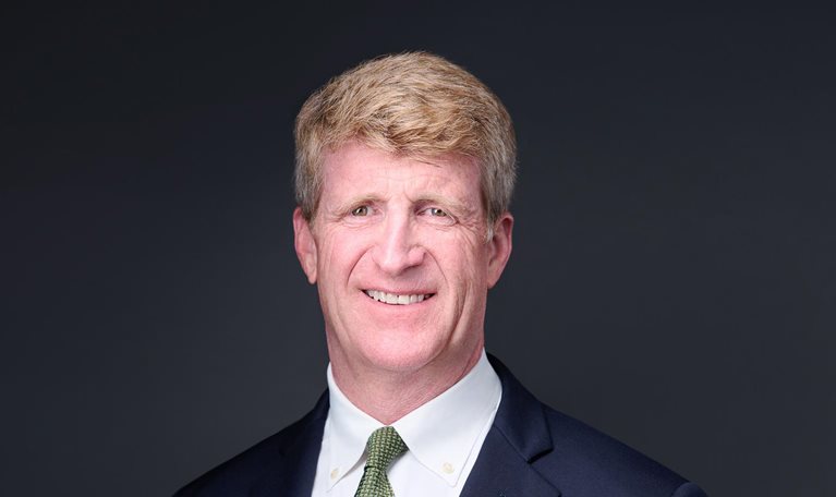 Patrick J. Kennedy, wearing a navy suit jacket, a green tie, and a smile, photographed against a dark background.