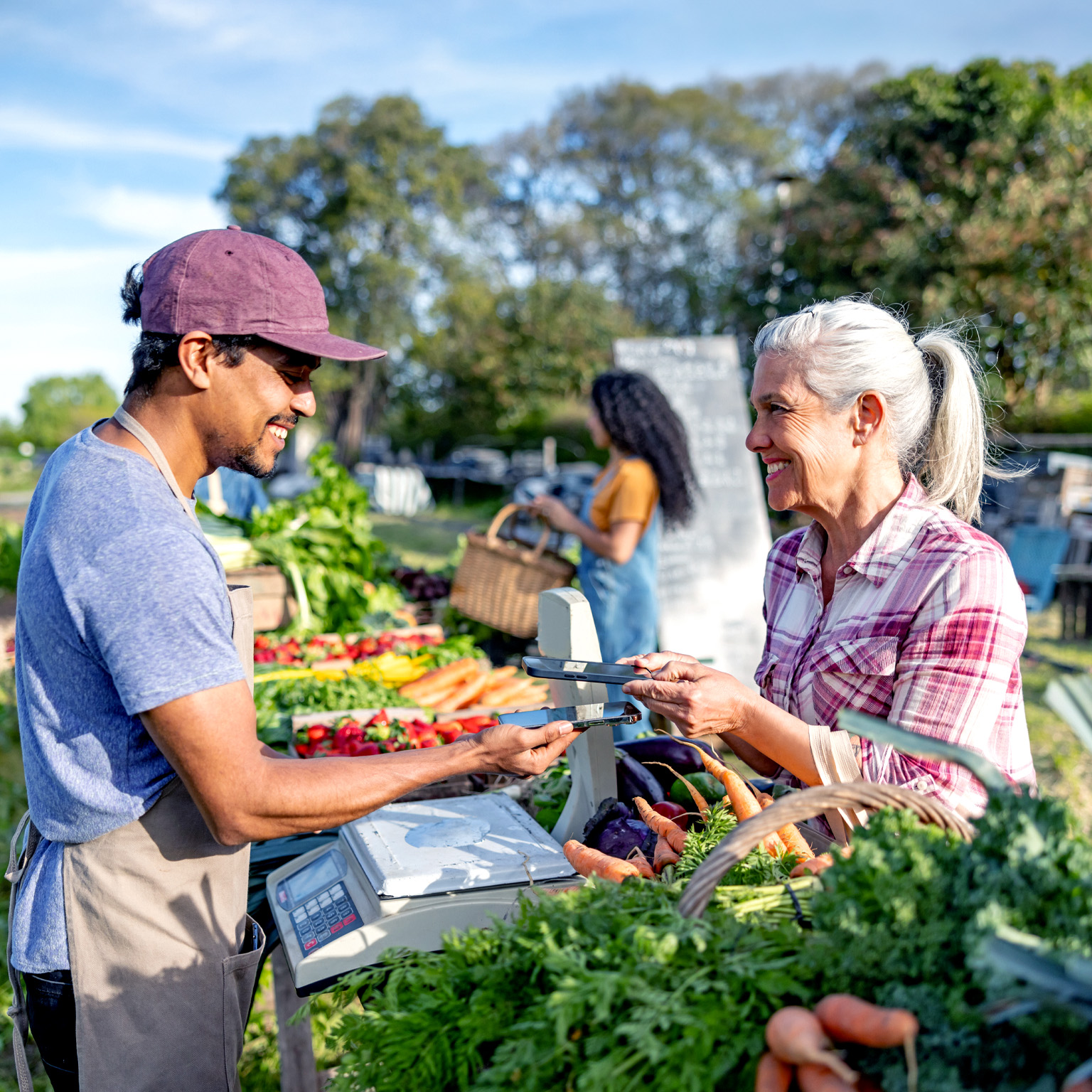 A vendor and a customer exchange a payment device across a produce stand filled with carrots, strawberries, and leafy greens at an outdoor market; another shopper stands in the background holding a basket.