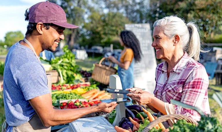 A vendor and a customer exchange a payment device across a produce stand filled with carrots, strawberries, and leafy greens at an outdoor market; another shopper stands in the background holding a basket.
