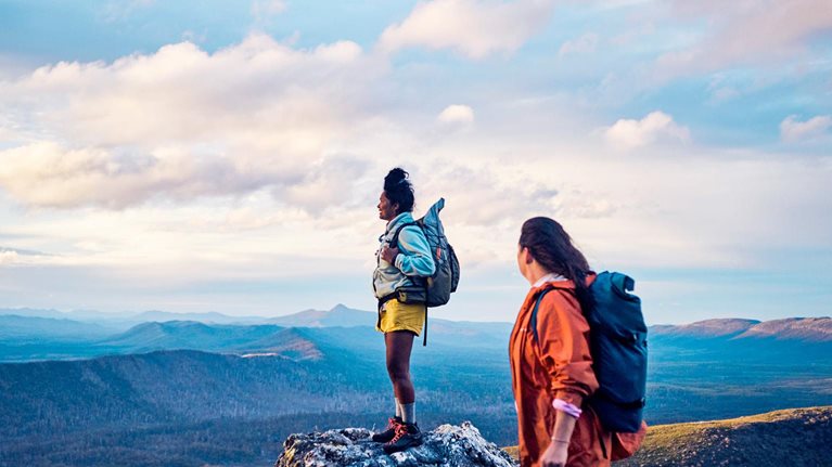 Two hikers stand on a rocky mountain peak, wearing backpacks and looking out over a vast, scenic landscape. The expansive view and soft light convey a sense of adventure, achievement, and connection with nature.
