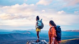 Two hikers stand on a rocky mountain peak, wearing backpacks and looking out over a vast, scenic landscape. The expansive view and soft light convey a sense of adventure, achievement, and connection with nature.
