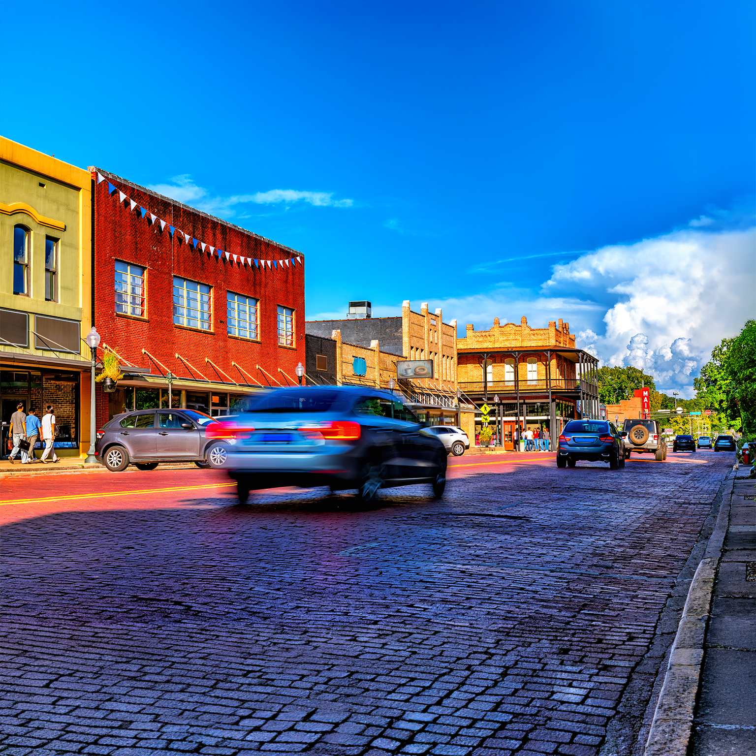 Cars and pedestrians are moving by on a busy main street in rural America with window-lined with shops and a cobblestone roadway