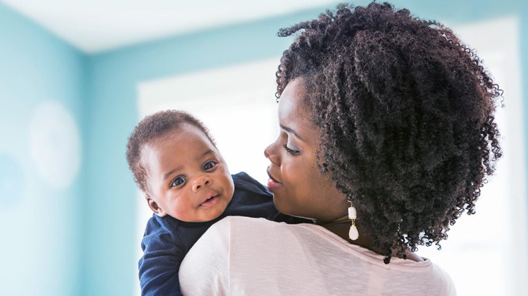 A Black mother with tightly curled ringlets holds her baby on her shoulder. The baby gazes at the viewer inquisitively as the mother looks at him thoughtfully.