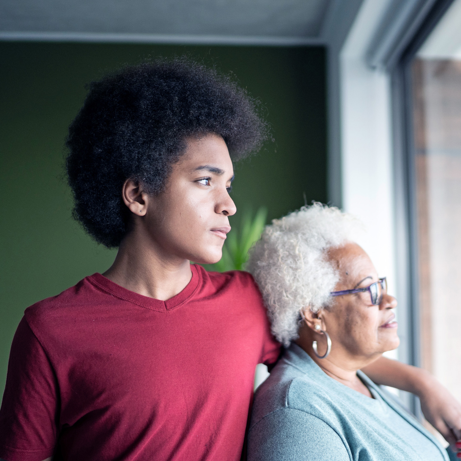 A young African American man stands beside his grandmother, his arm resting gently on her shoulder as they both gaze thoughtfully out of a window. 