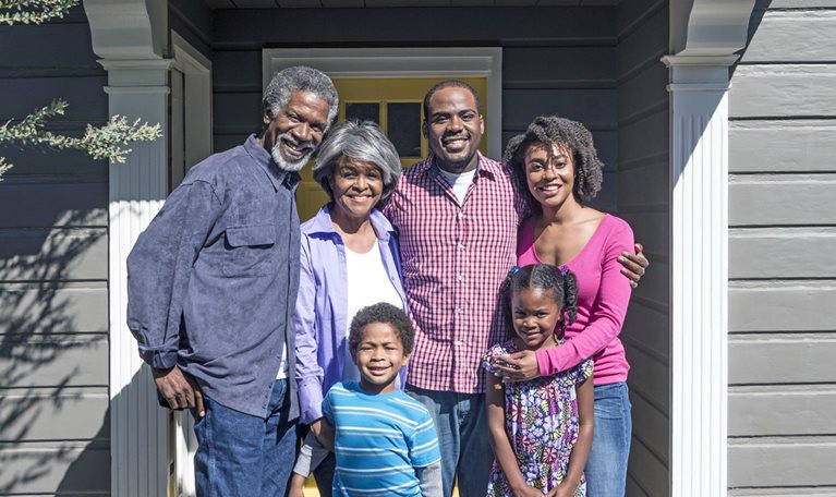 Three generations of a Black family pose together and smile outside their home.
